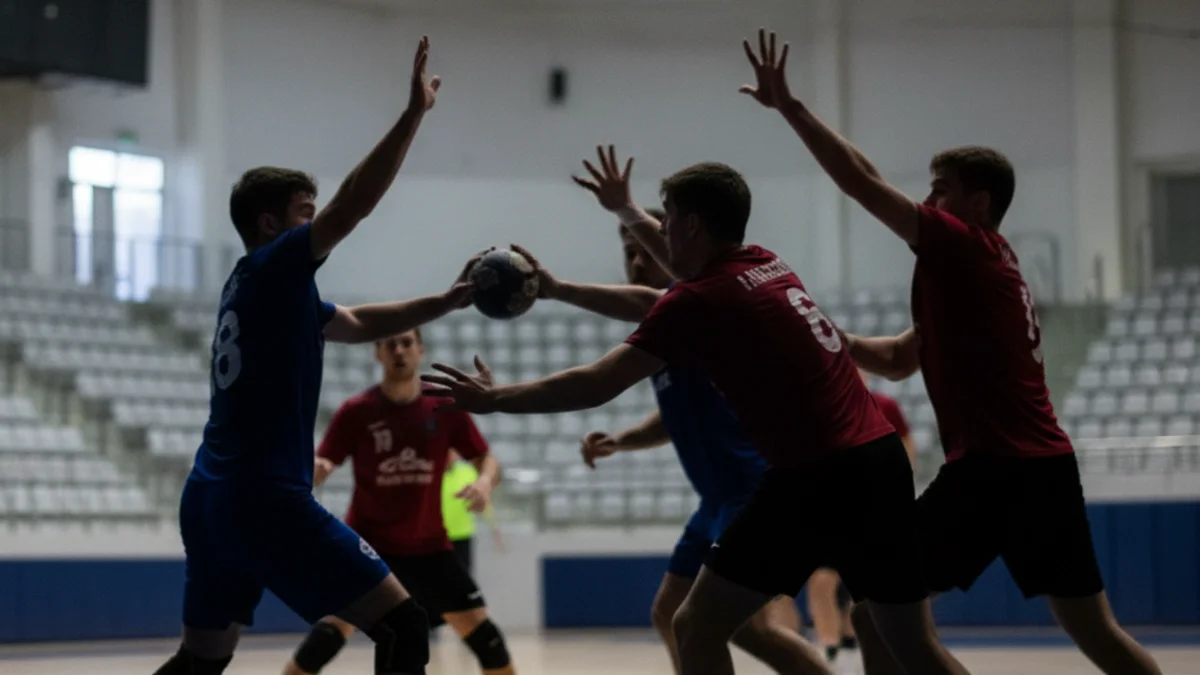 Imatge genèrica d'un partit d'handbol en un pavelló esportiu, amb jugadors en acció i la pilota en l'aire.