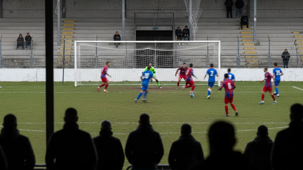 Vista d'un camp de futbol local durant un partit de lliga territorial, amb figures borroses de jugadors.