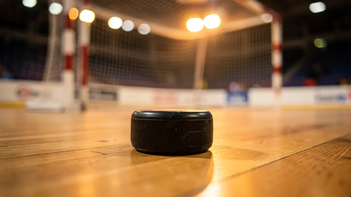 Primer plano de un disco de hockey patines sobre parquet, con la portería y luces del estadio desenfocados al fondo.