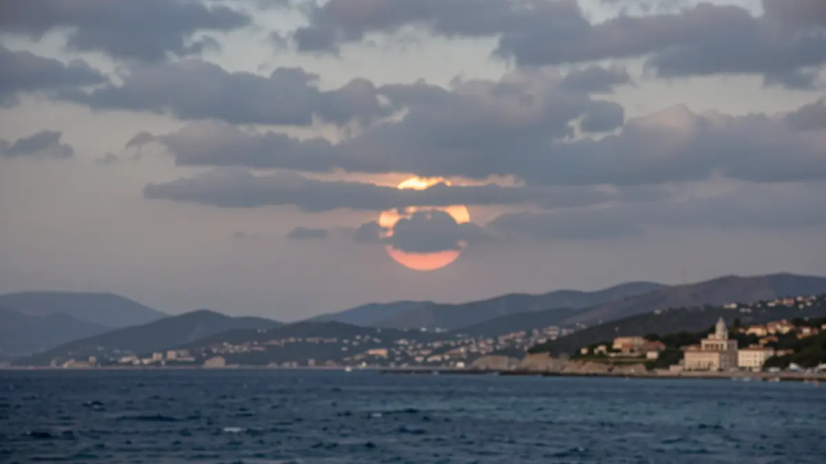 Partially cloudy sky over a Mediterranean coastline with hills in the background during a solar eclipse.