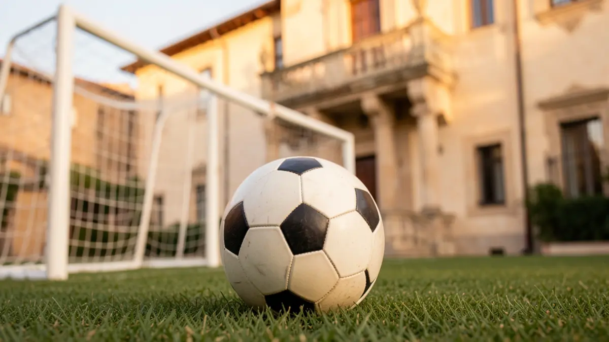 Generic image of a soccer ball on grass with a blurred goal net in the background and a Mediterranean town hall facade in the soft focus distance.