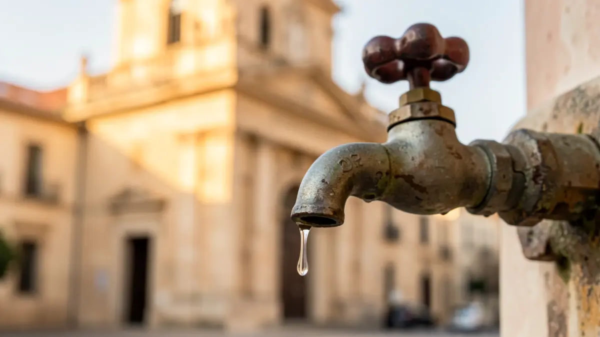 Imatge genèrica d'una aixeta amb una gota d'aigua i, al fons, la façana d'un ajuntament mediterrani.