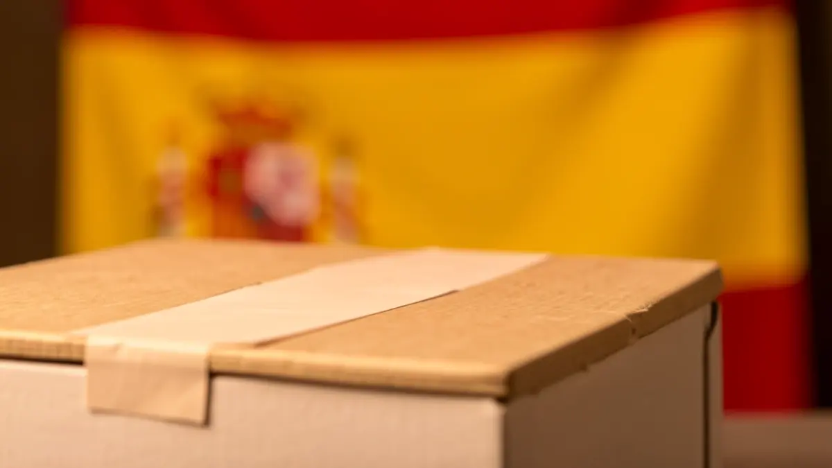 Imatge genèrica d'una urna electoral espanyola amb una bandera espanyola difuminada al fons.